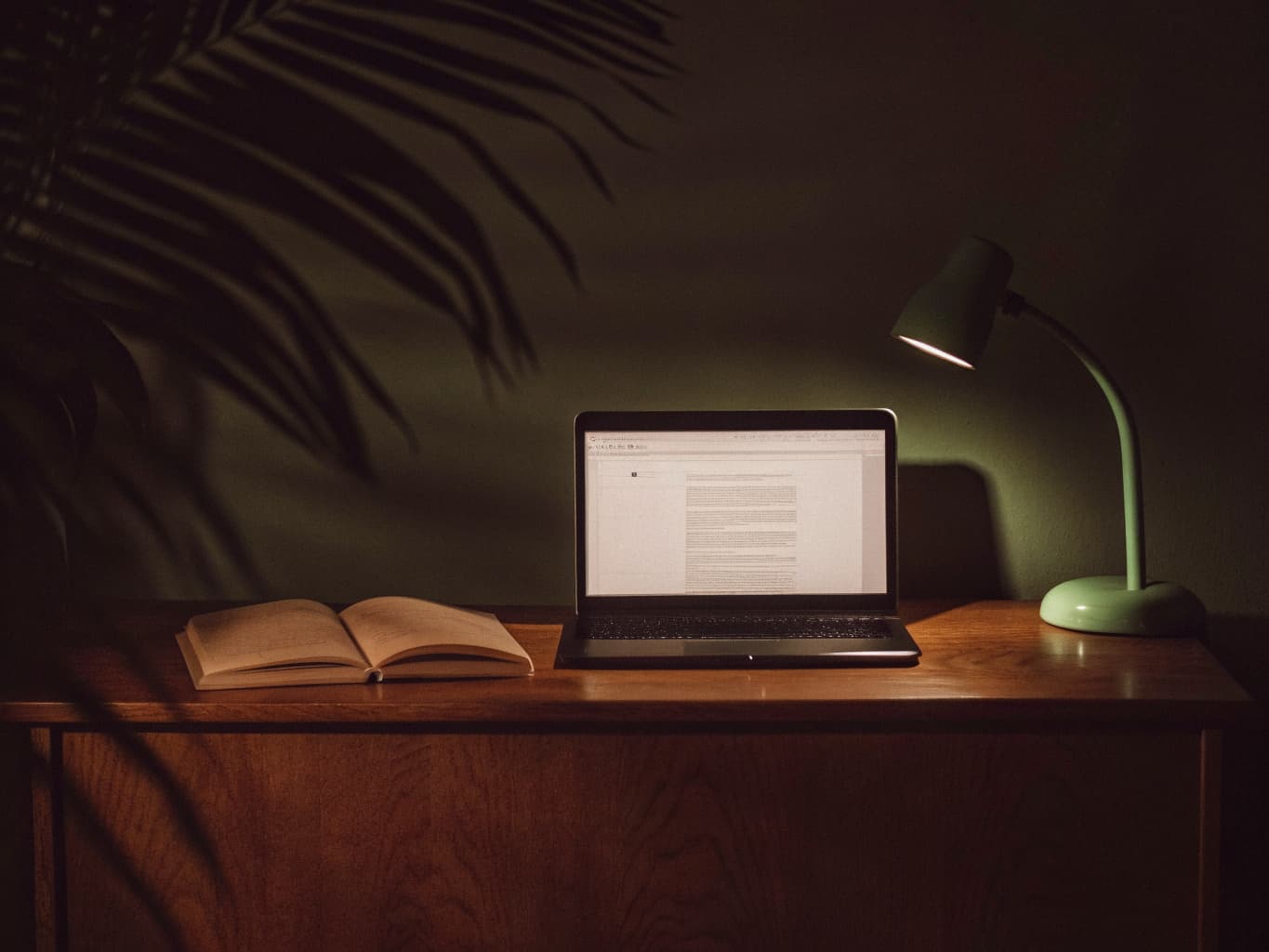 An open notebook next to a laptop showing a blog article being drafted, on a warm wood desk with a mint-green lamp glow